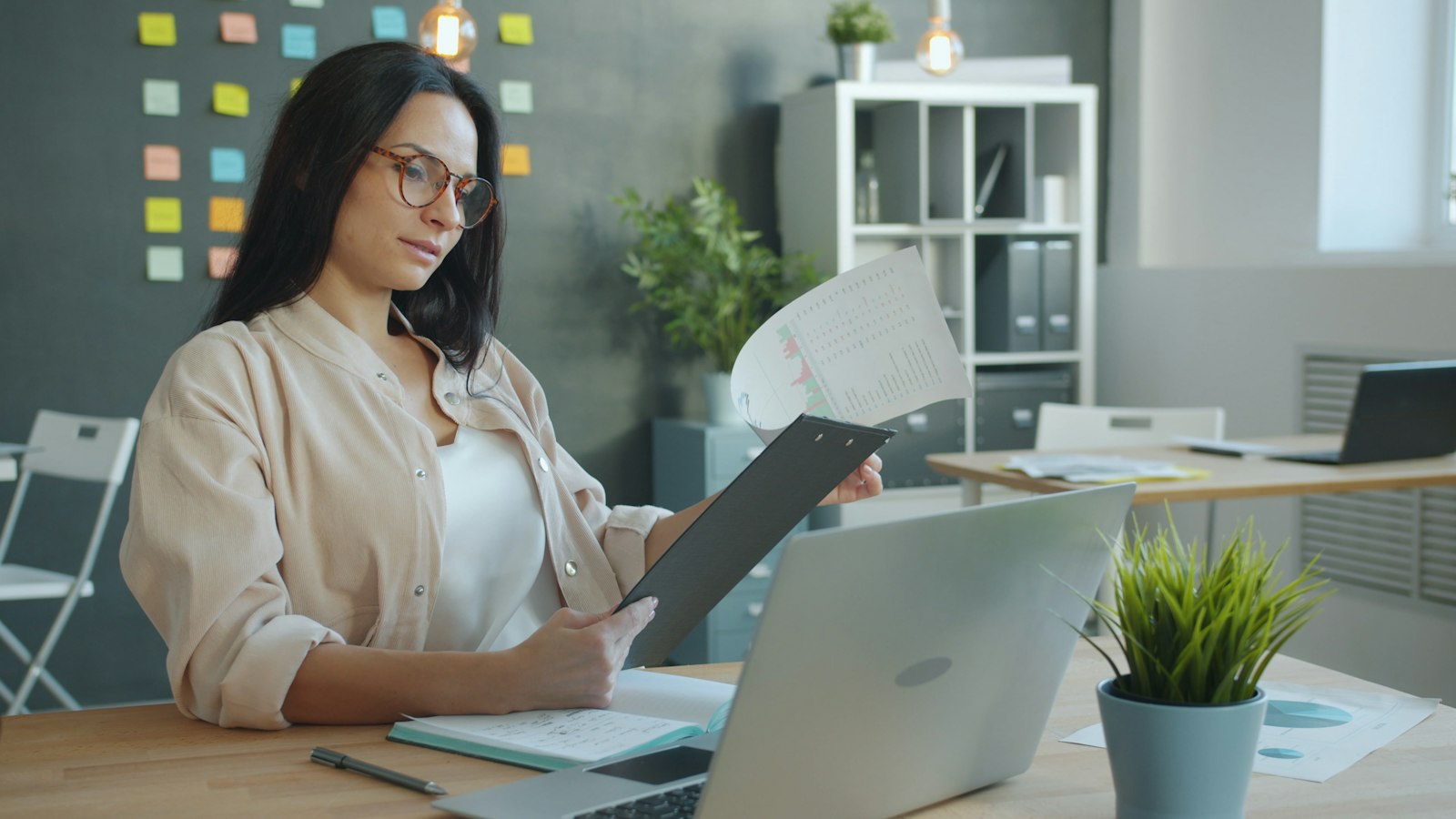 A woman in glasses reviews a printed report on a clipboard at her office desk, with an open laptop and a notebook in front of her and binders on a shelf behind.
