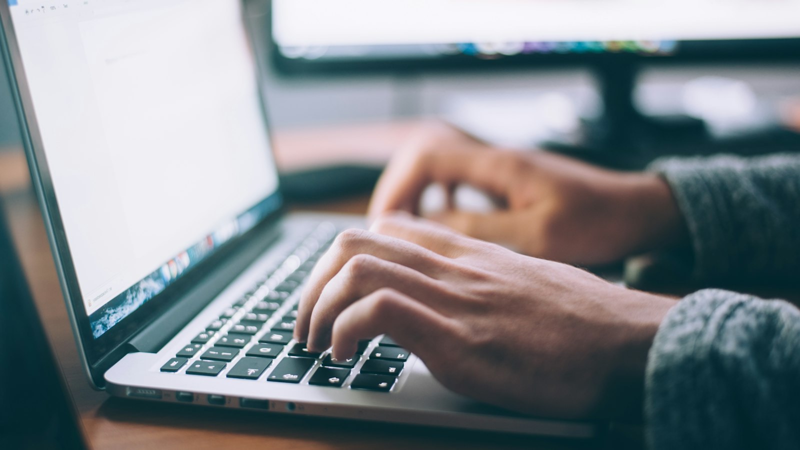 Hands typing on a laptop keyboard at a desk, a second monitor glowing out of focus behind it.