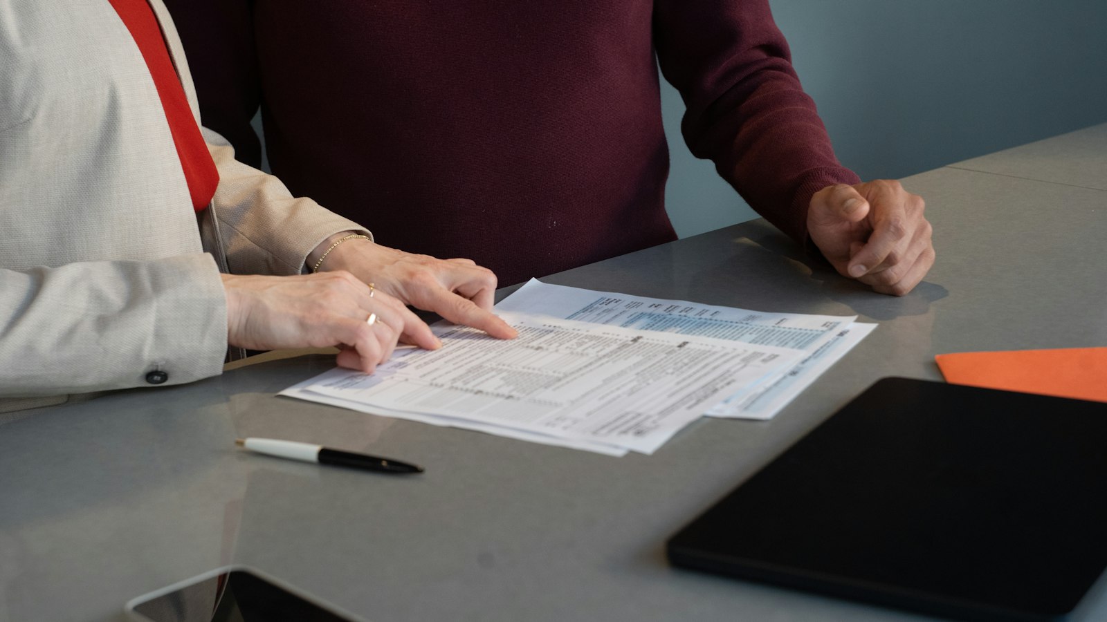 Two colleagues at a desk going through a printed sheet of figures together, a pen and tablet beside them.