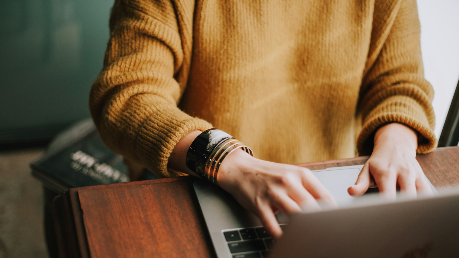 A person in a mustard knitted jumper typing on a laptop at a wooden desk in warm daylight.
